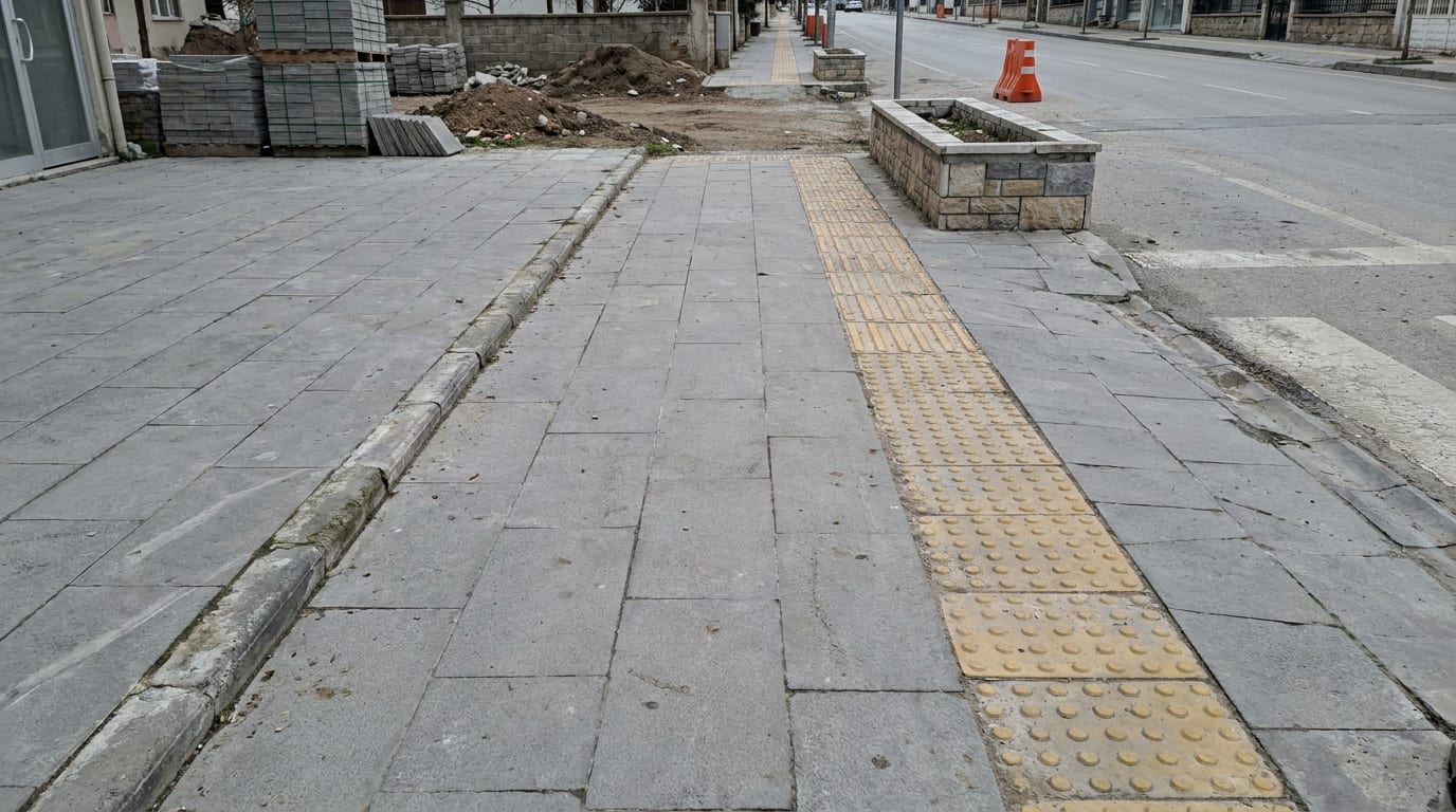 Sokak boyunca gri beton plaka kaldırım; bordür hattı ve geçişte hissedilebilir şerit